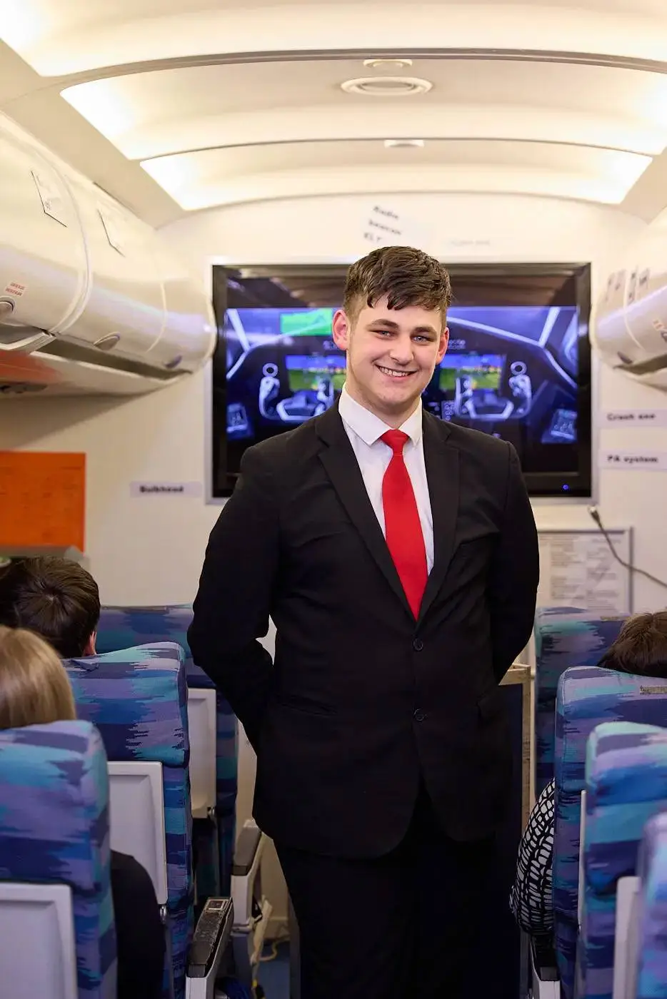 A man in a suit and red tie stands in a train carriage, smiling at the camera. Behind him, a screen displays safety information.