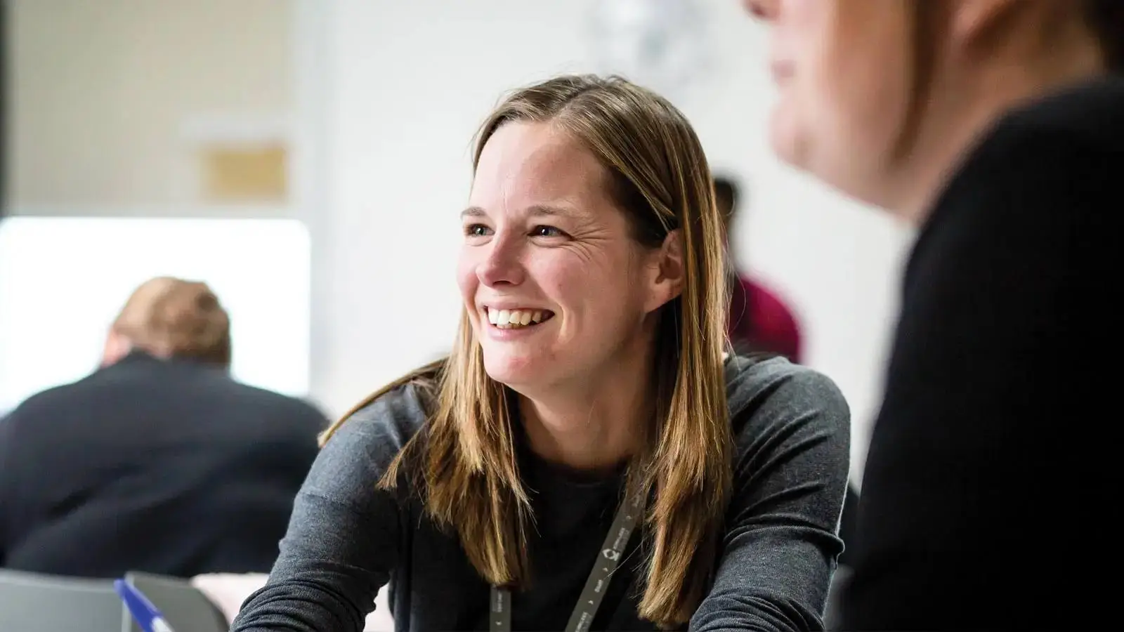 A woman smiling and sitting at a table in a classroom.