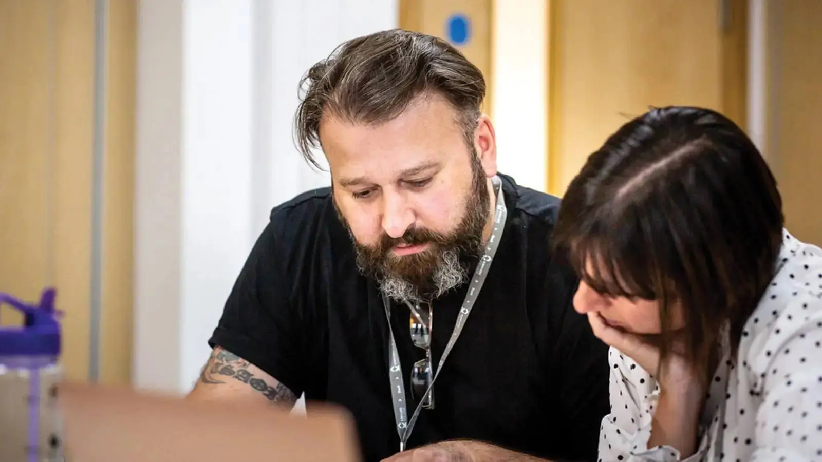 Two people in a classroom, one man and one woman, looking at a laptop screen. The man has a beard and is wearing a black t-shirt. The woman has her hand on her chin.