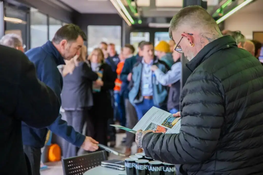 A man in a black puffer jacket reading a brochure at a college event with people in the background.