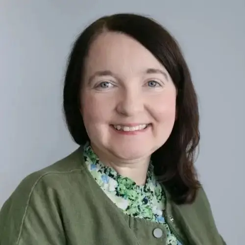 A woman in a green cardigan and floral blouse smiles against a grey background. She has short dark hair and blue eyes.