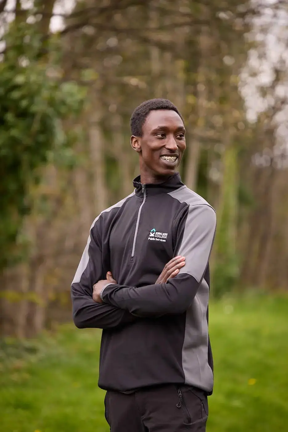 A man in a black and grey tracksuit stands in a field with trees behind him, smiling and looking to the side.