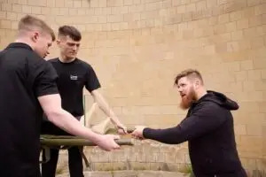 Three men in black shirts arm wrestling over a green prosthetic arm in front of a beige brick wall.