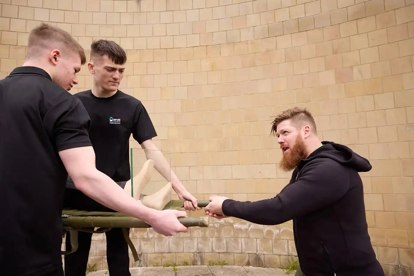 Three men in black shirts arm wrestling over a green prosthetic arm in front of a beige brick wall.
