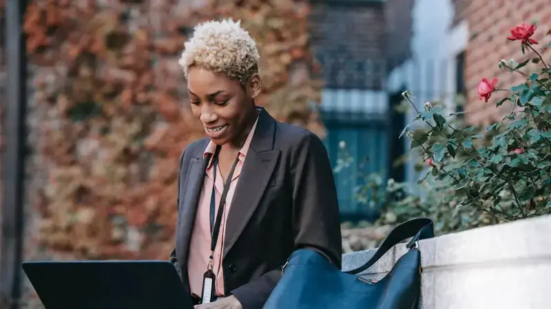 A person in a suit using a laptop outdoors, surrounded by autumn leaves and flowers, suggesting a blend of nature and modern education.