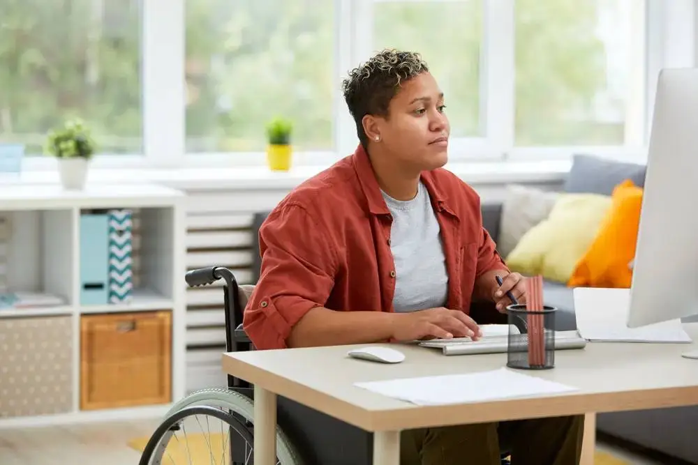 A student in a wheelchair using a computer at a desk in a bright room with plants and a sofa.