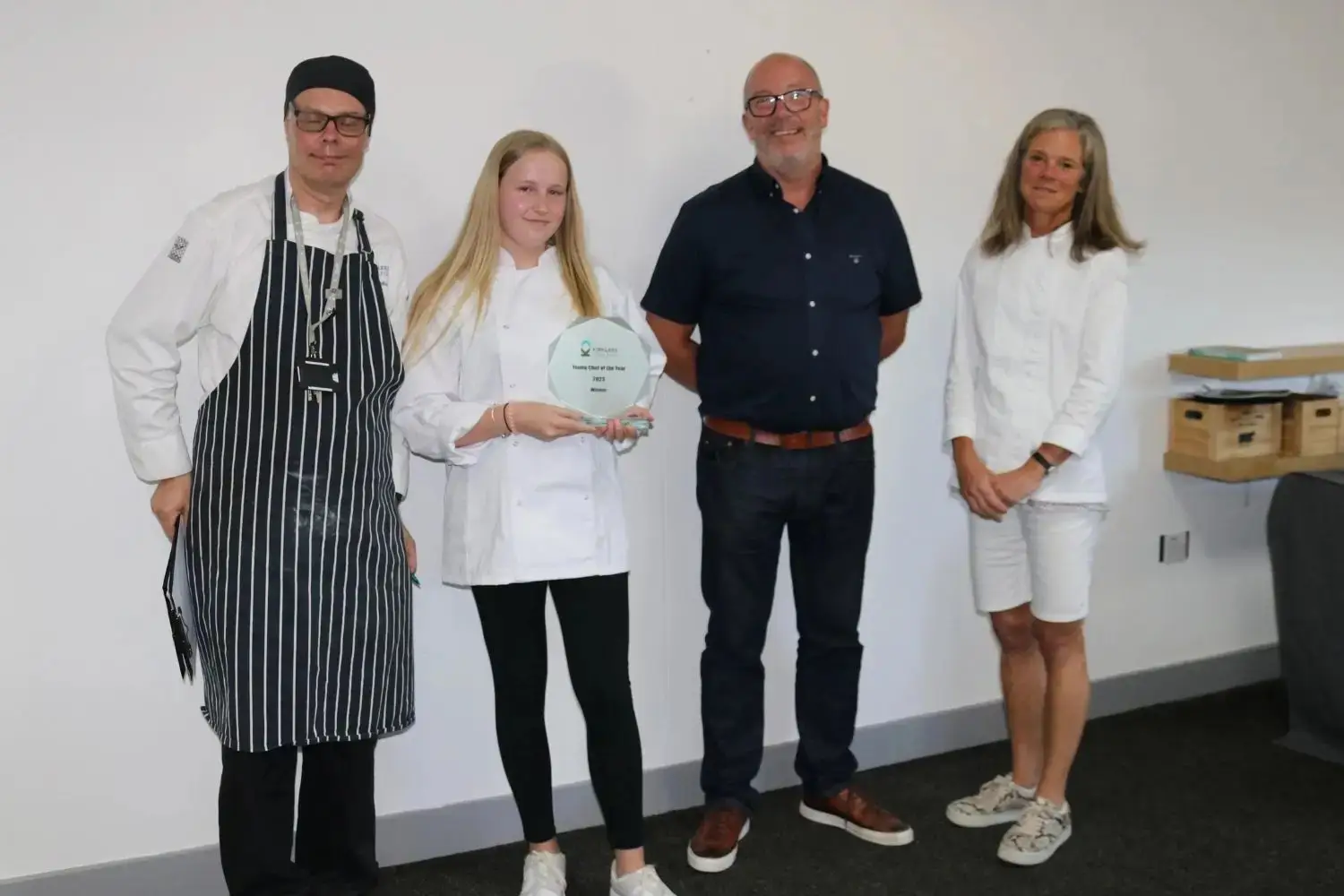 A young woman in a chef's coat holds a plaque, flanked by three adults, celebrating an achievement in culinary education.