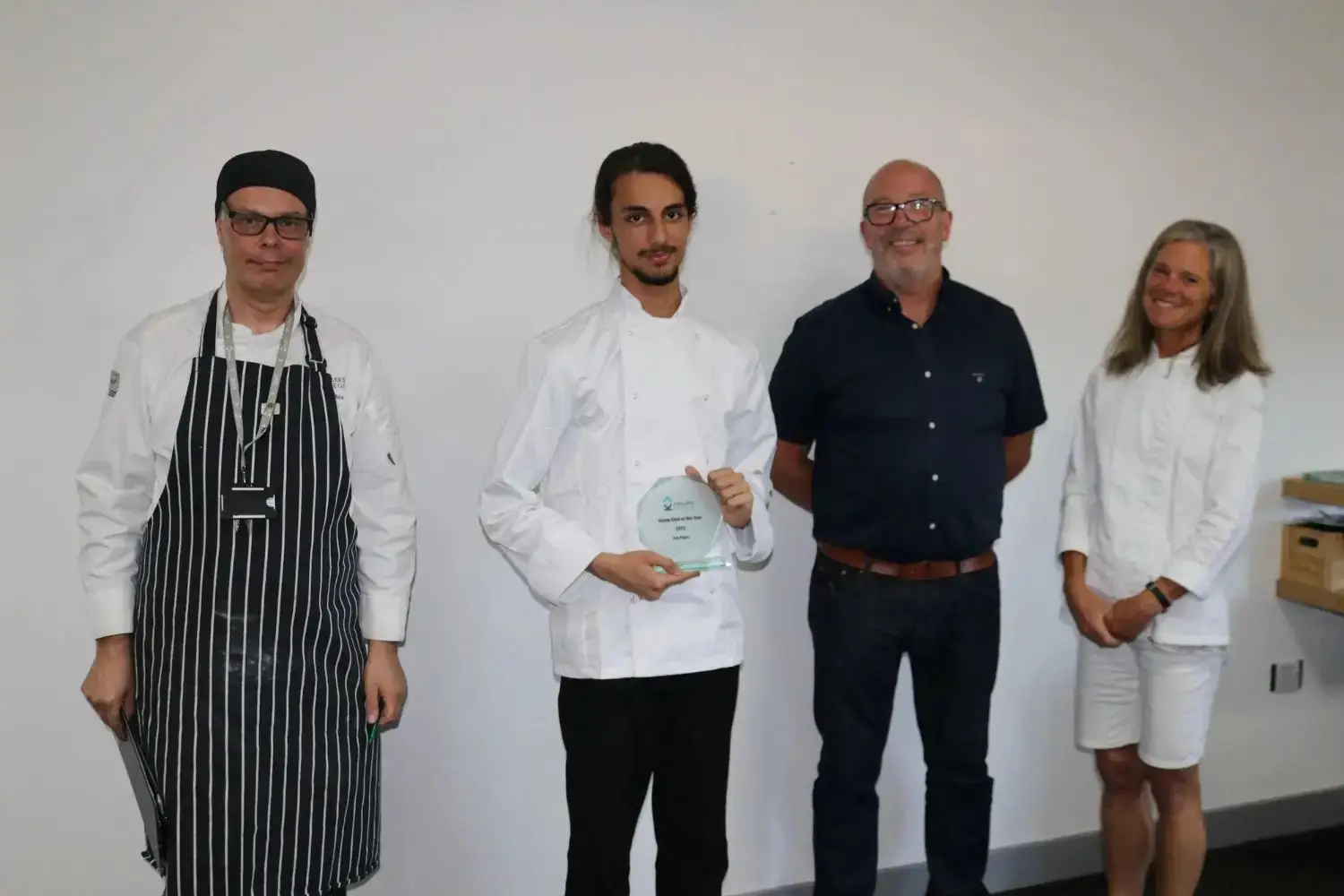 A chef in a white uniform holding a glass award stands with three colleagues in a college setting.