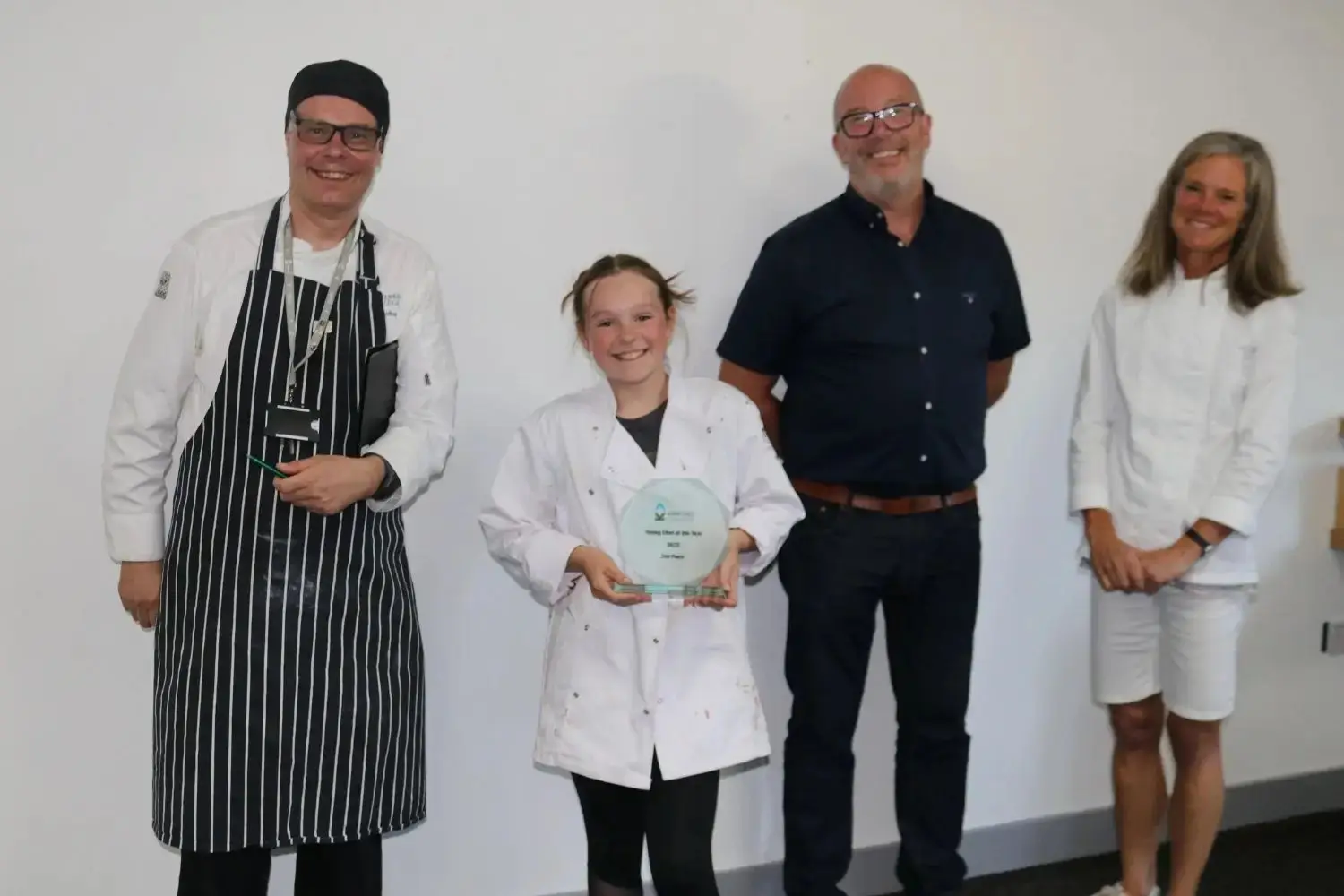 A young student in a white lab coat holds a blue award plaque, flanked by three adults in chef and academic attire, smiling in a college setting.