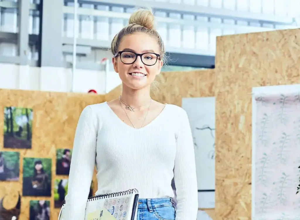 A smiling student in glasses holds a notebook, standing in front of a corkboard with photos and sketches in a modern college setting.