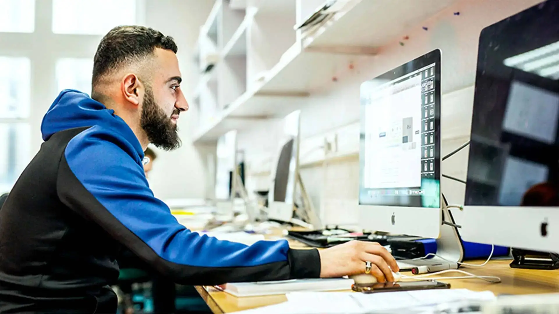 A student in a blue and black hoodie working on a desktop computer in a brightly lit college computer lab.