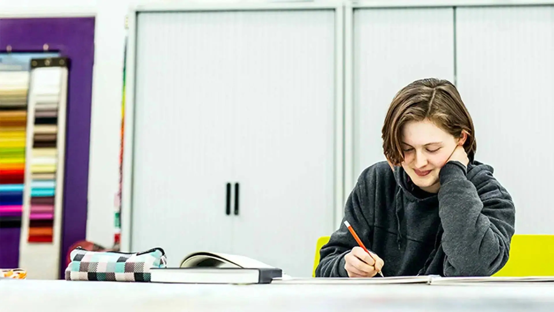 A student in a grey hoodie writing in a notebook at a white desk in a classroom with a colour swatch board.