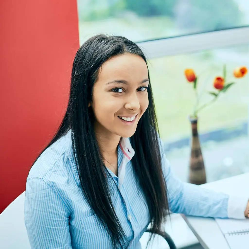 A smiling woman in a blue shirt sits at a desk with a laptop and a vase of flowers, looking out a window.