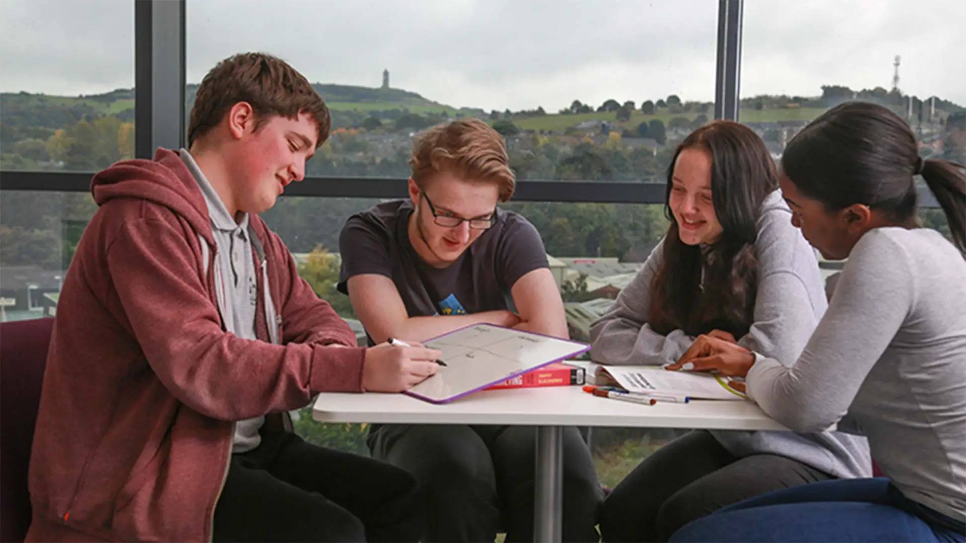 Four students in casual wear sitting around a table, engaged in a group study session with notebooks and pens, overlooking a scenic landscape through large windows.