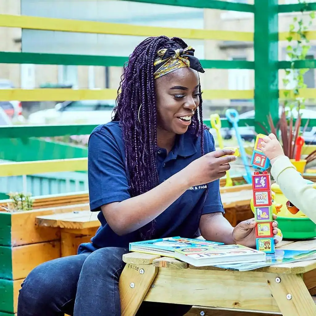 A woman in a blue polo shirt sits at a wooden table, smiling and engaging with a child using colourful stacking blocks and educational cards.