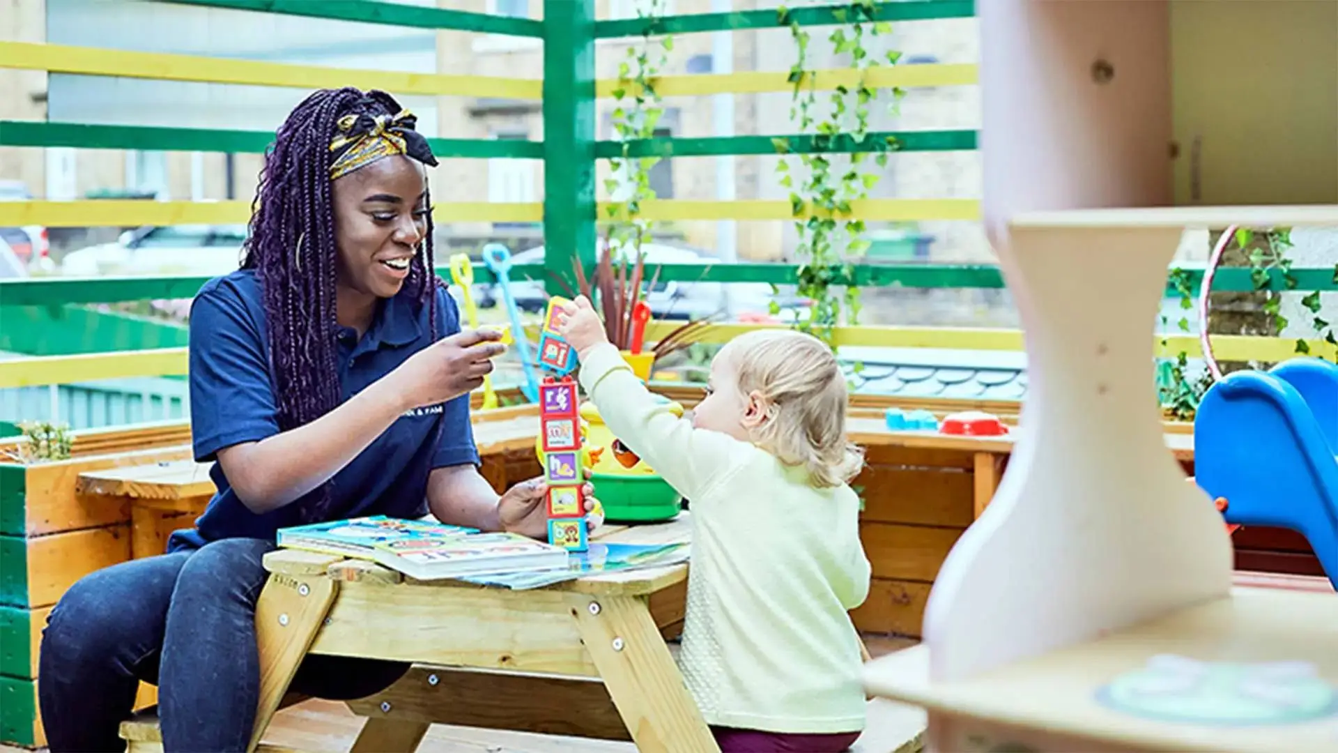 A woman and a child playing with a stacking toy at a wooden table in a brightly lit room with green and yellow railings.