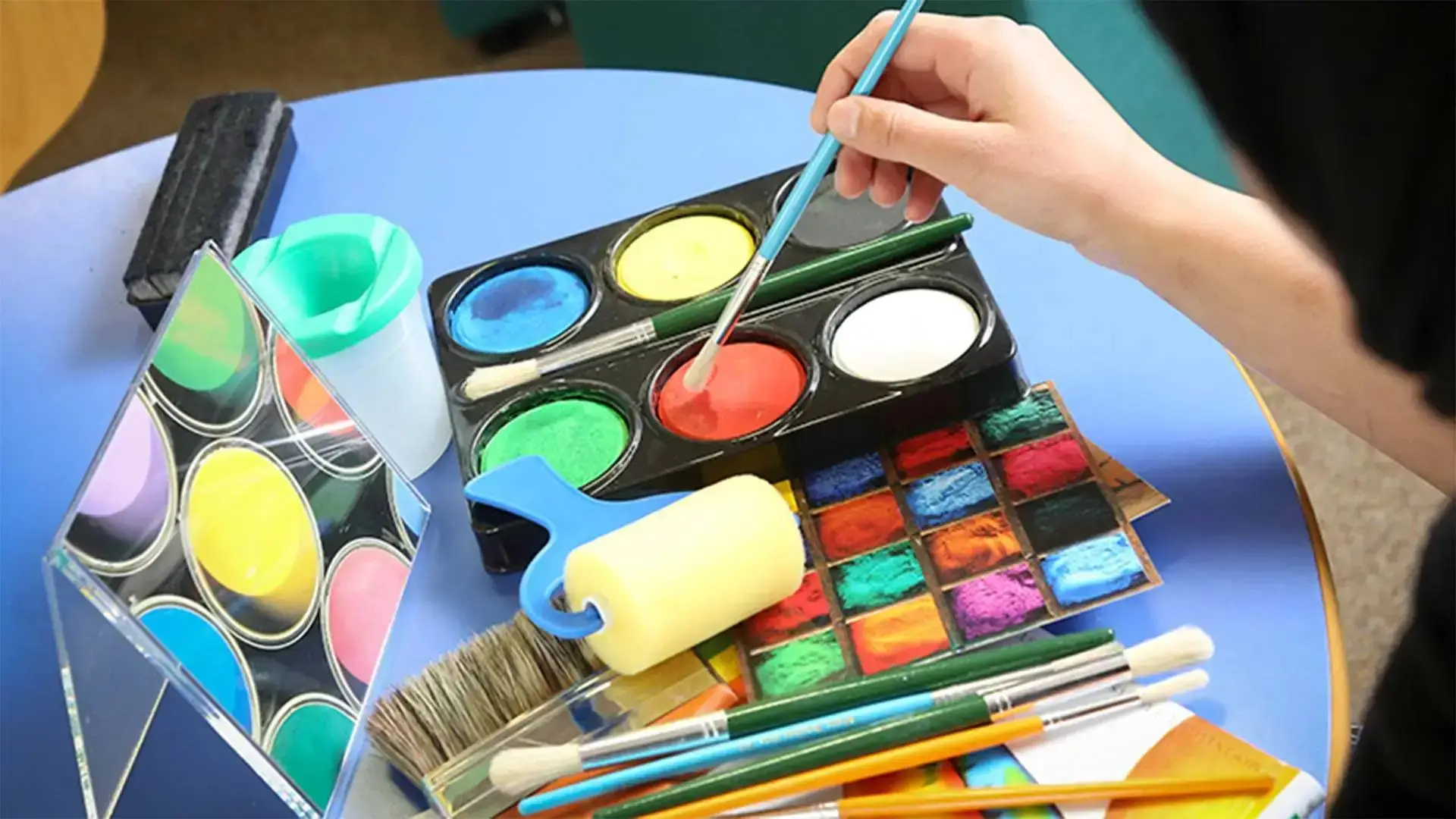 A person paints with a brush on a palette of vibrant watercolours, surrounded by brushes and paint tubes on a blue table.