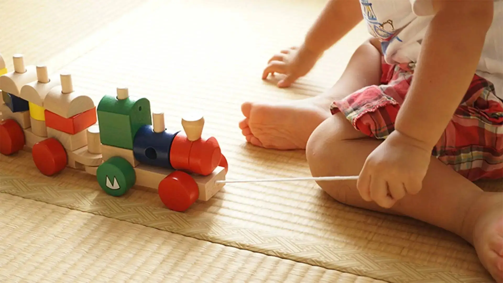 A child sitting on a mat pulling a colourful wooden train with a string.