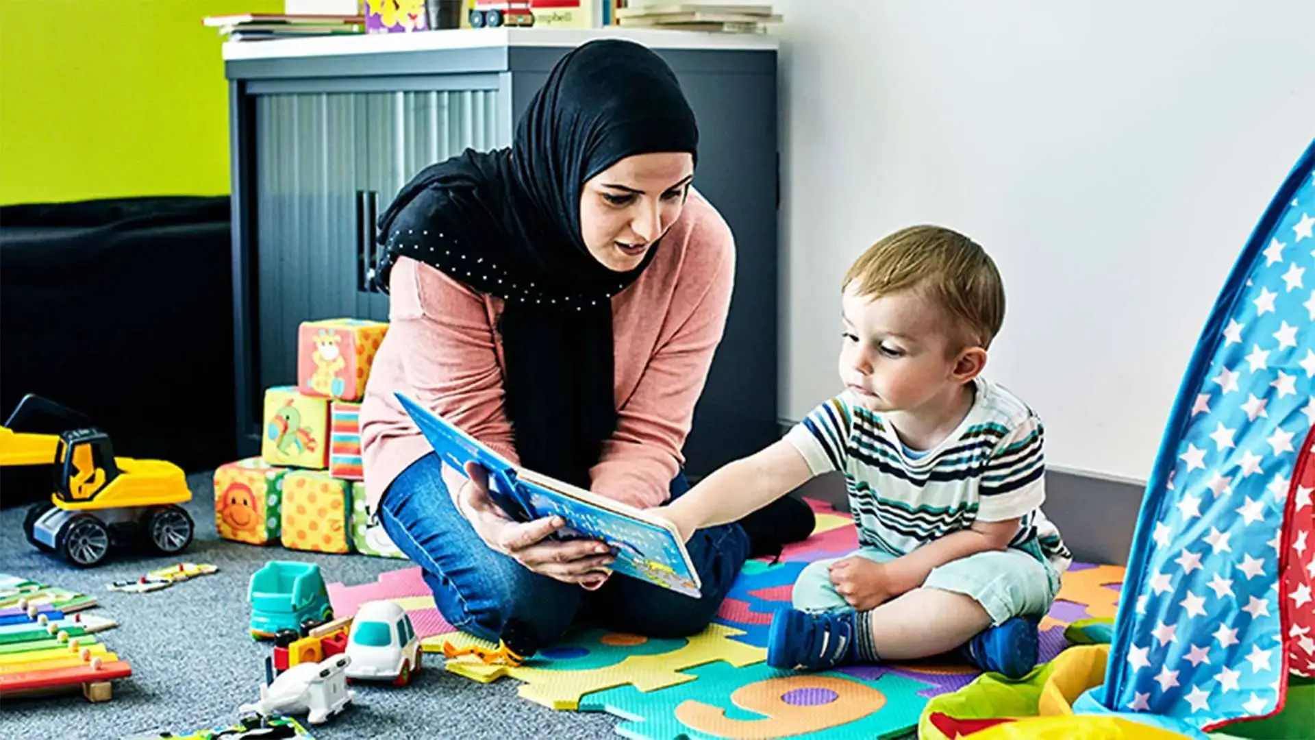 A woman reading a book to a child on a colourful play mat surrounded by toys.