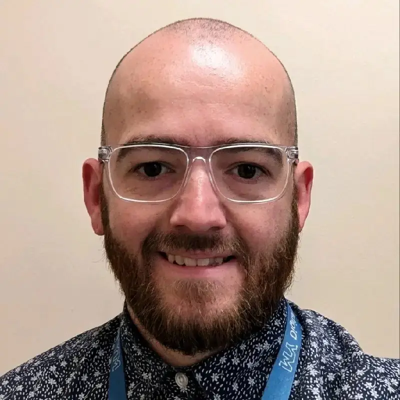 A bald man with glasses and a beard smiles, wearing a patterned shirt and a blue lanyard. Background is plain.