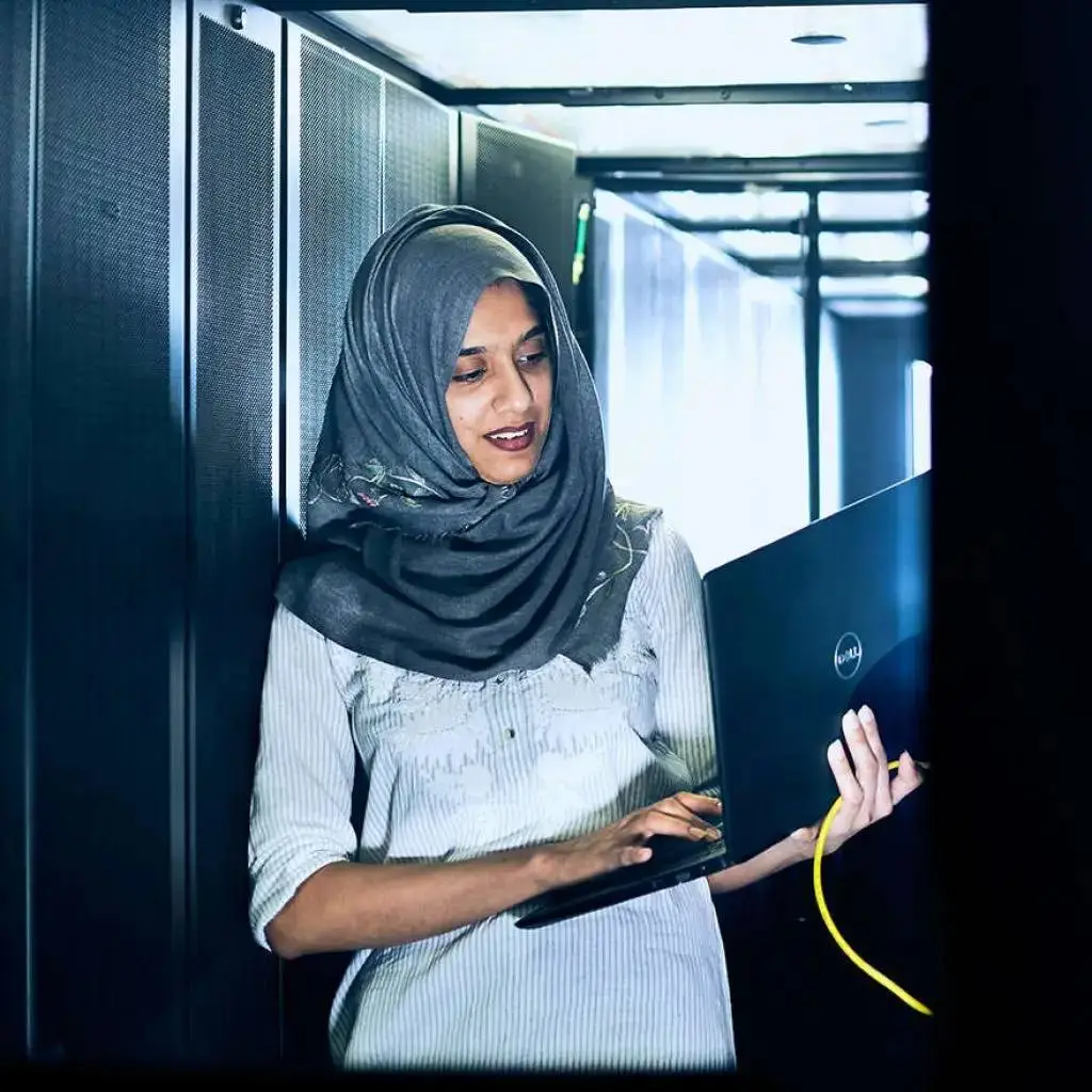 A woman in a grey headscarf using a laptop in front of server racks in a data centre.