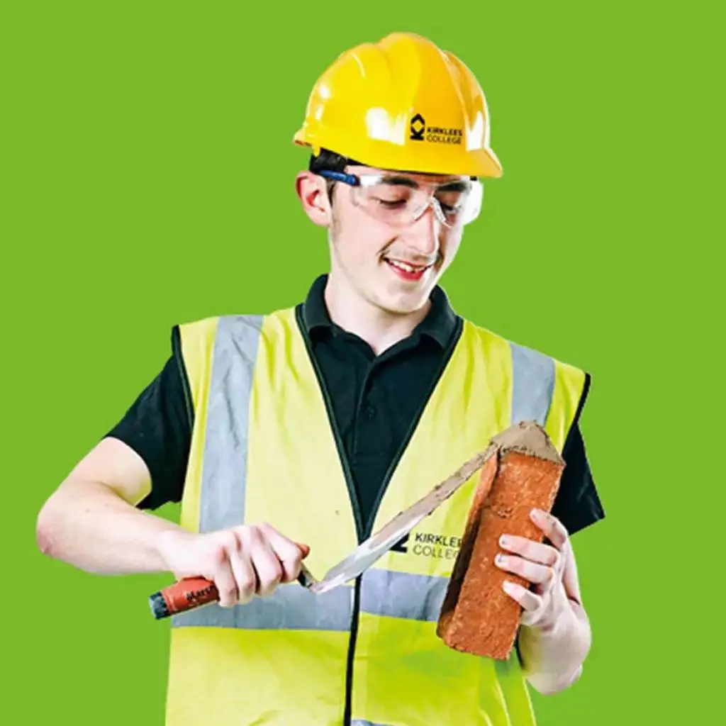 A student in a high-vis vest and hard hat cutting a brick with a saw, smiling, against a green background. Kirklees