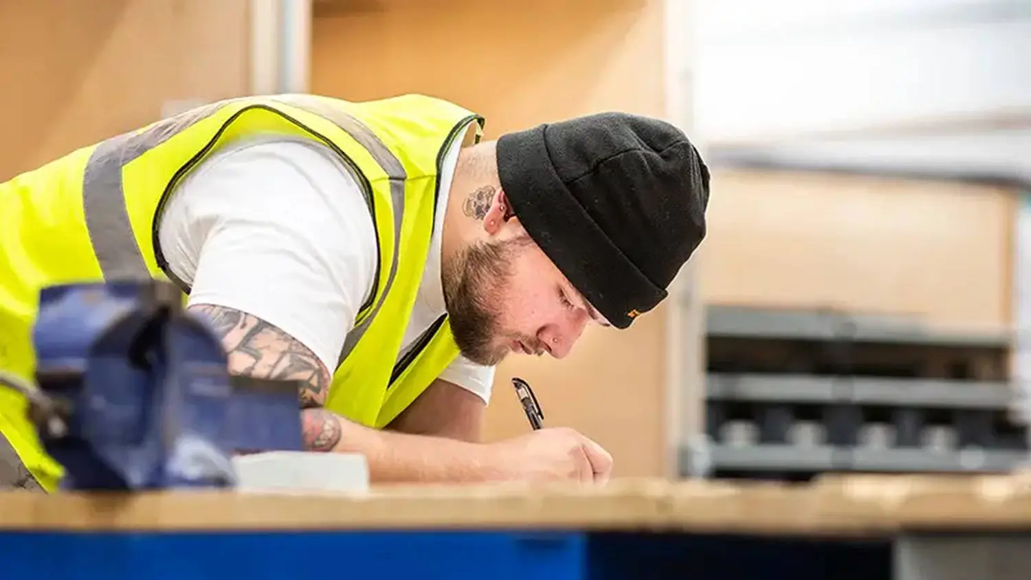 A man in a high-visibility vest and beanie writing on a wooden surface in a workshop.