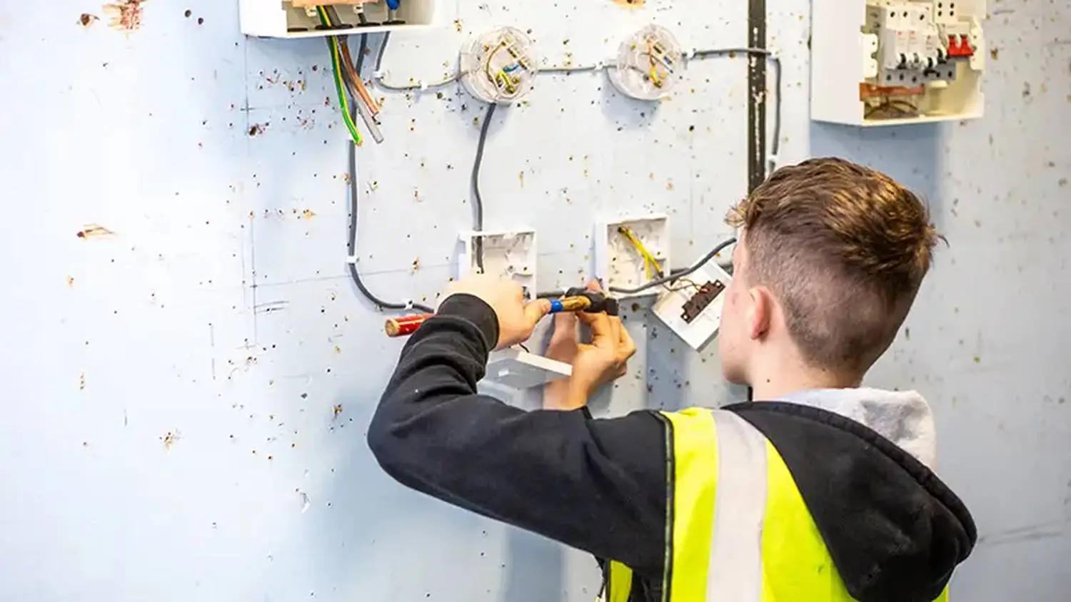 A person in a high-visibility vest working on electrical wiring in a room with exposed plaster and a consumer unit.
