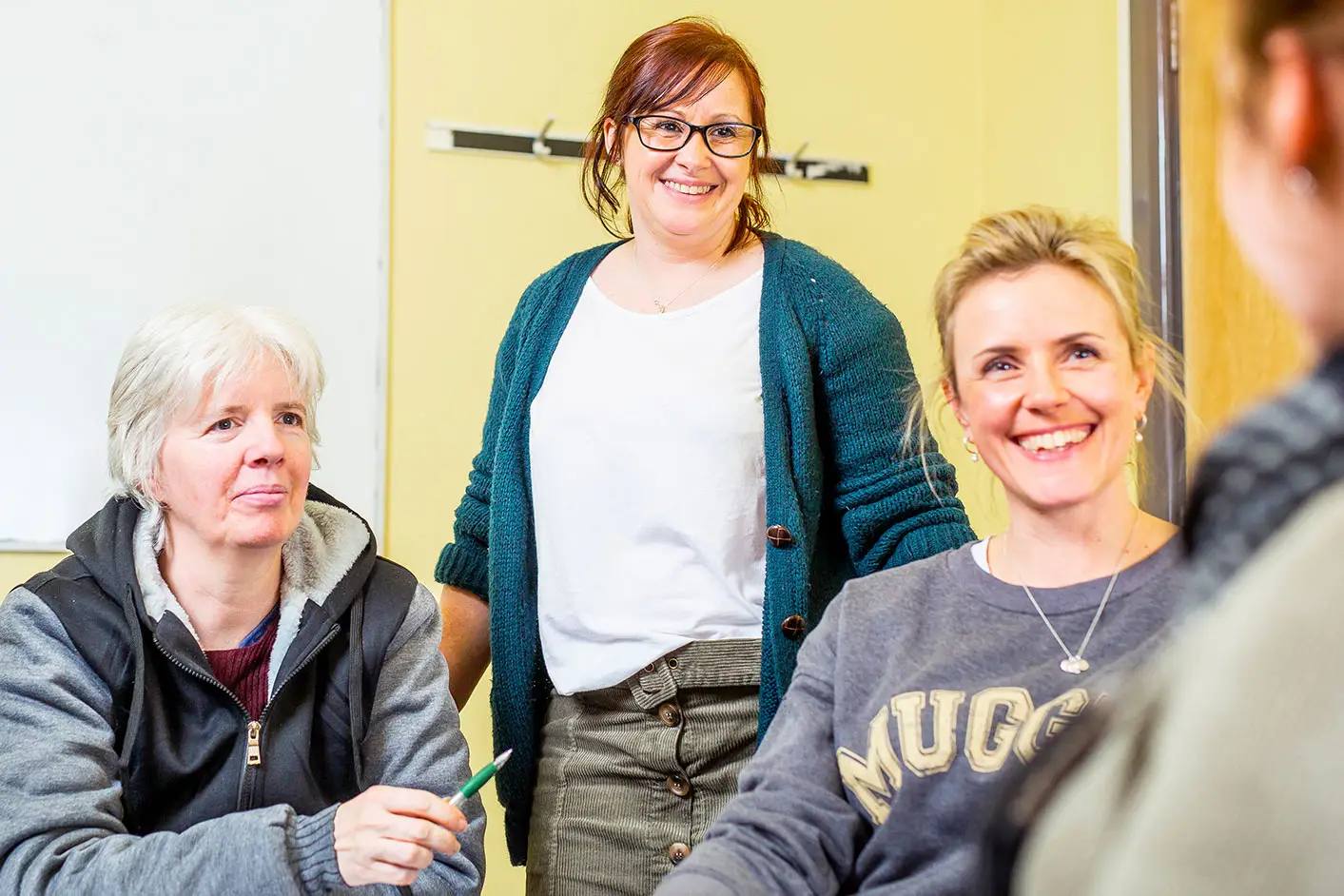 Three women smiling in a classroom setting, engaged in discussion. One stands, holding a pen, while the others sit attentively.