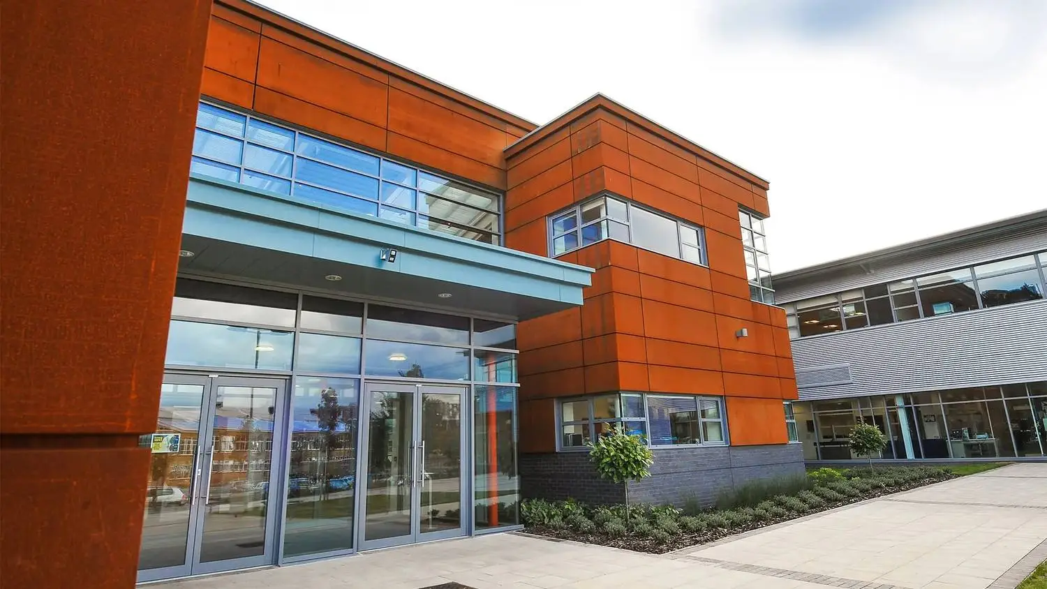 Modern college building with orange and glass facade, entrance with sliding doors, and landscaped pathway under a partly cloudy sky.