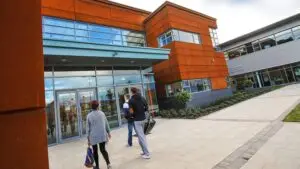 People walking towards a modern college building with glass doors and orange cladding, surrounded by landscaped pathways.