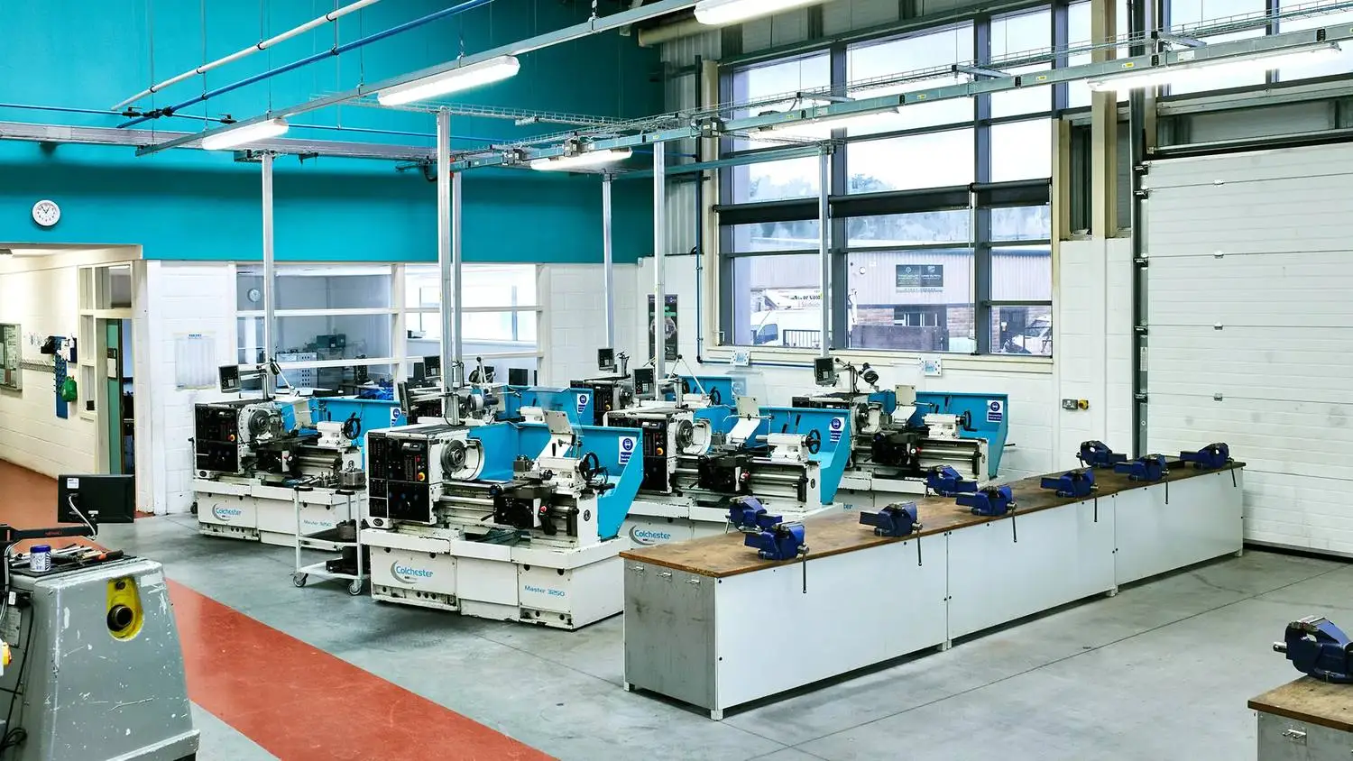 A college workshop with multiple blue and white lathes on wooden benches, under fluorescent lights, with large windows and a red and grey floor.
