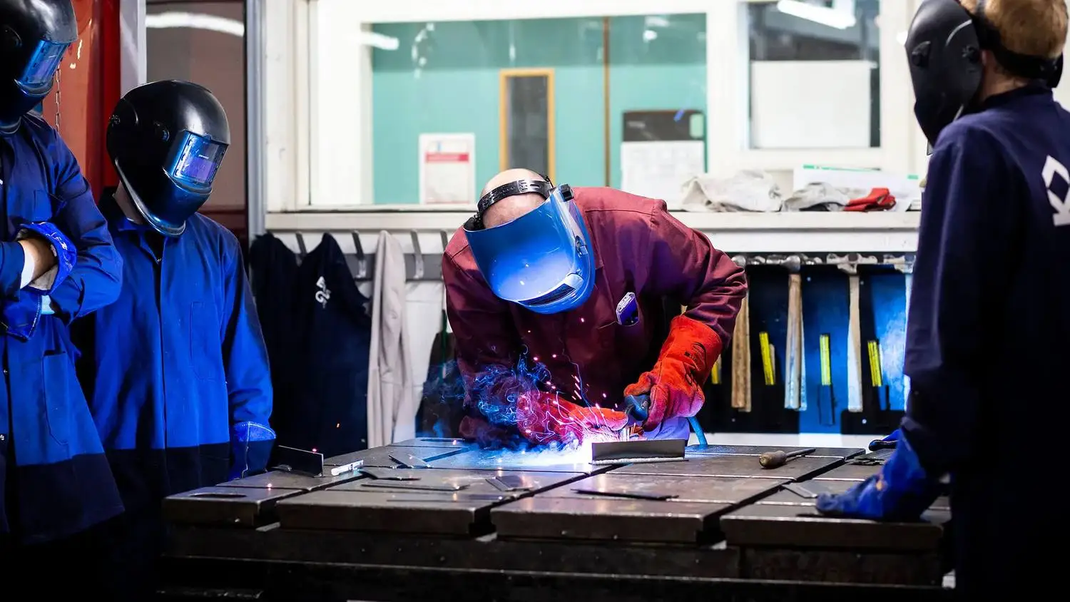 Students in protective gear welding metal in a workshop, learning practical skills in a hands-on educational setting.