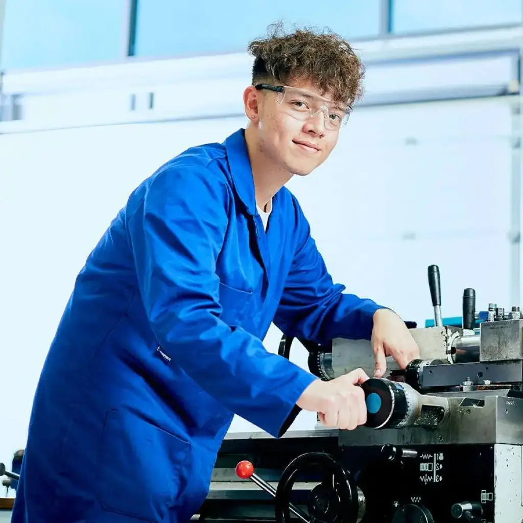 A student in a blue coverall and safety glasses operates a lathe machine in a well-lit workshop, focused on precision engineering tasks.