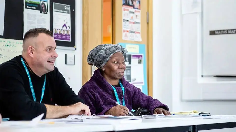 Two people in a classroom, one writing, both wearing lanyards, with educational posters on the walls.