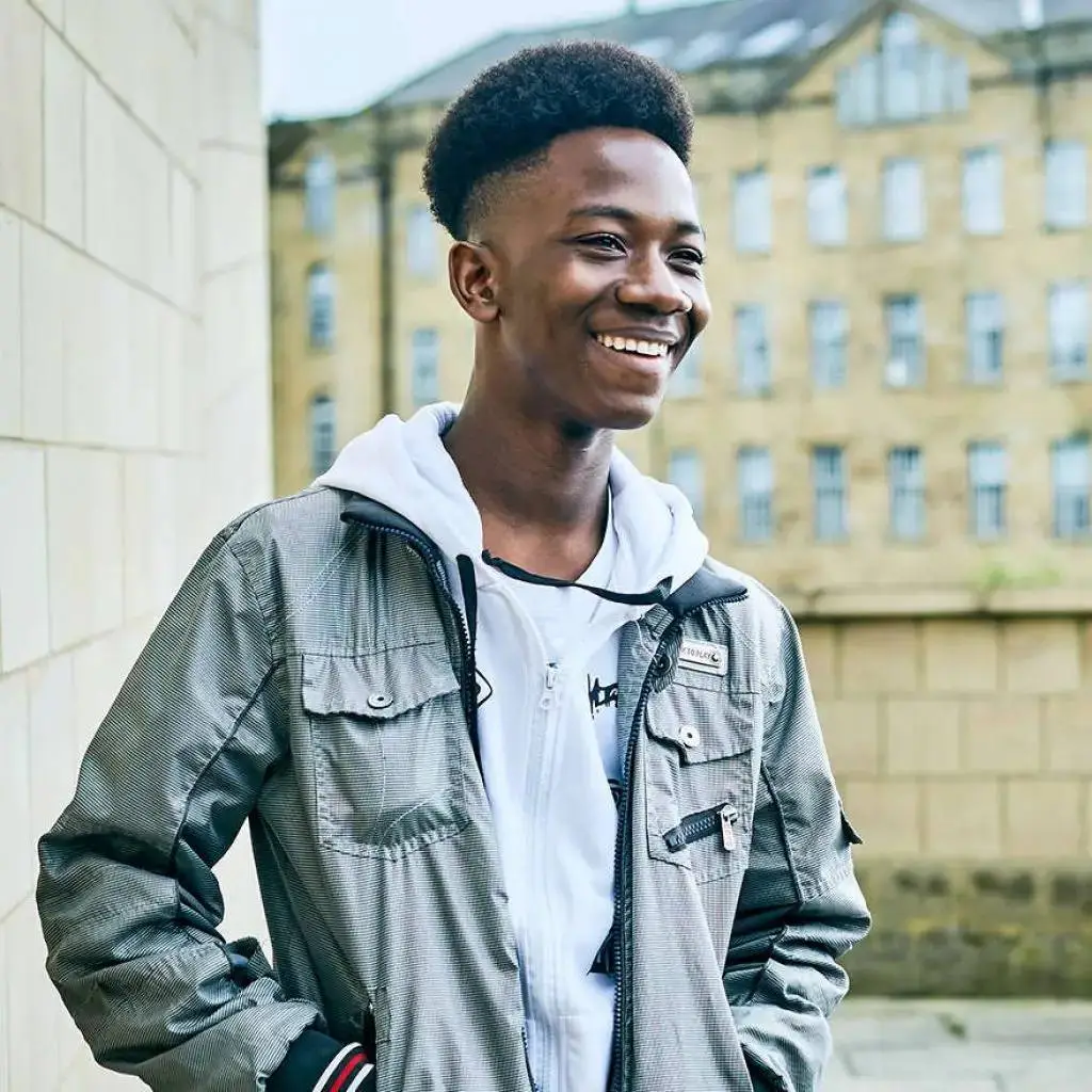 A young man in a grey jacket smiling in front of a stone building.