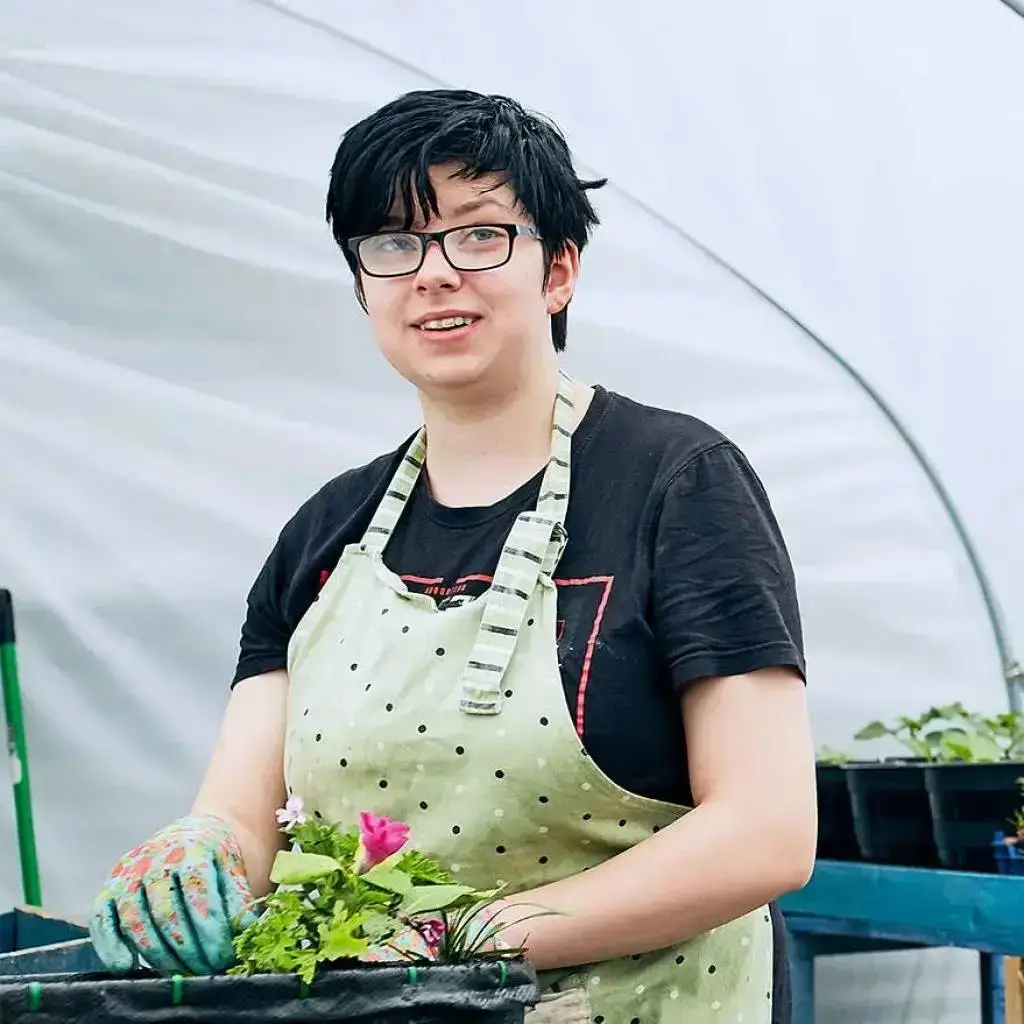 A person in a polka dot apron and floral gloves stands in a greenhouse, smiling and holding a tray of plants.