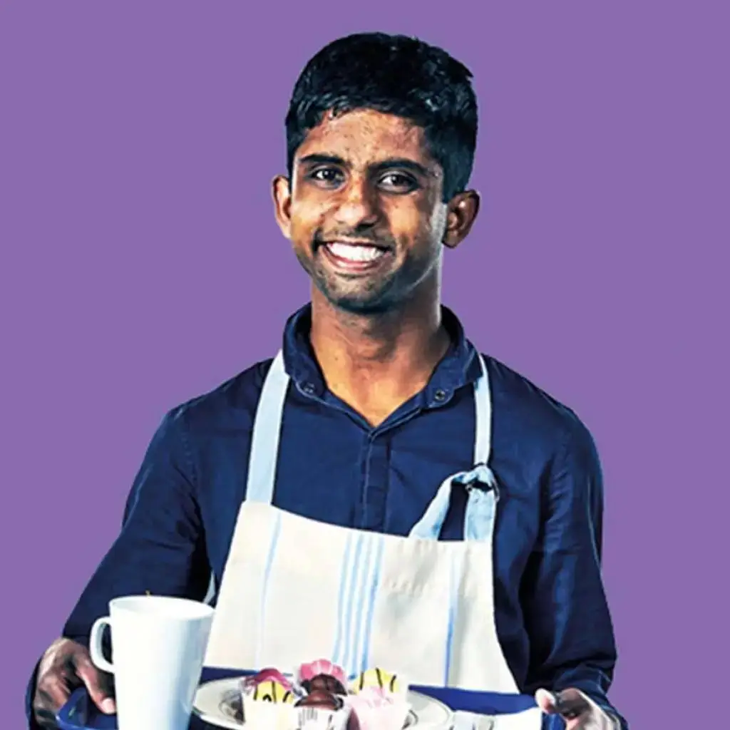A man in a blue shirt and white apron holds a tray with cupcakes and a mug, smiling against a purple background. Educational theme.