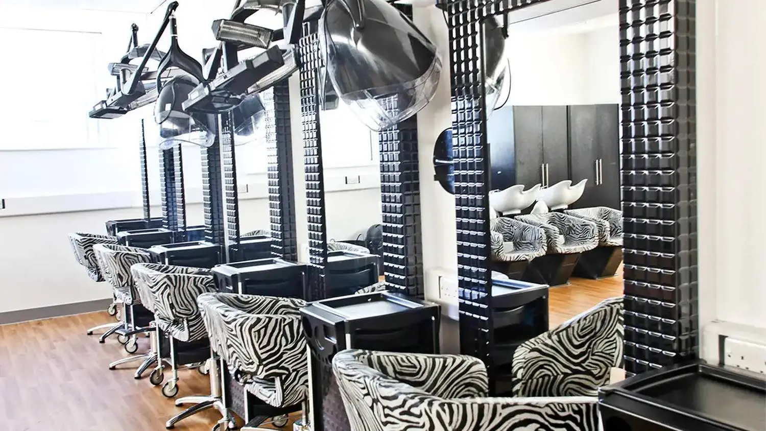 A modern hair salon with black and white zebra-patterned chairs, hooded dryers, and sleek black stations, reflecting a stylish and contemporary educational environment.