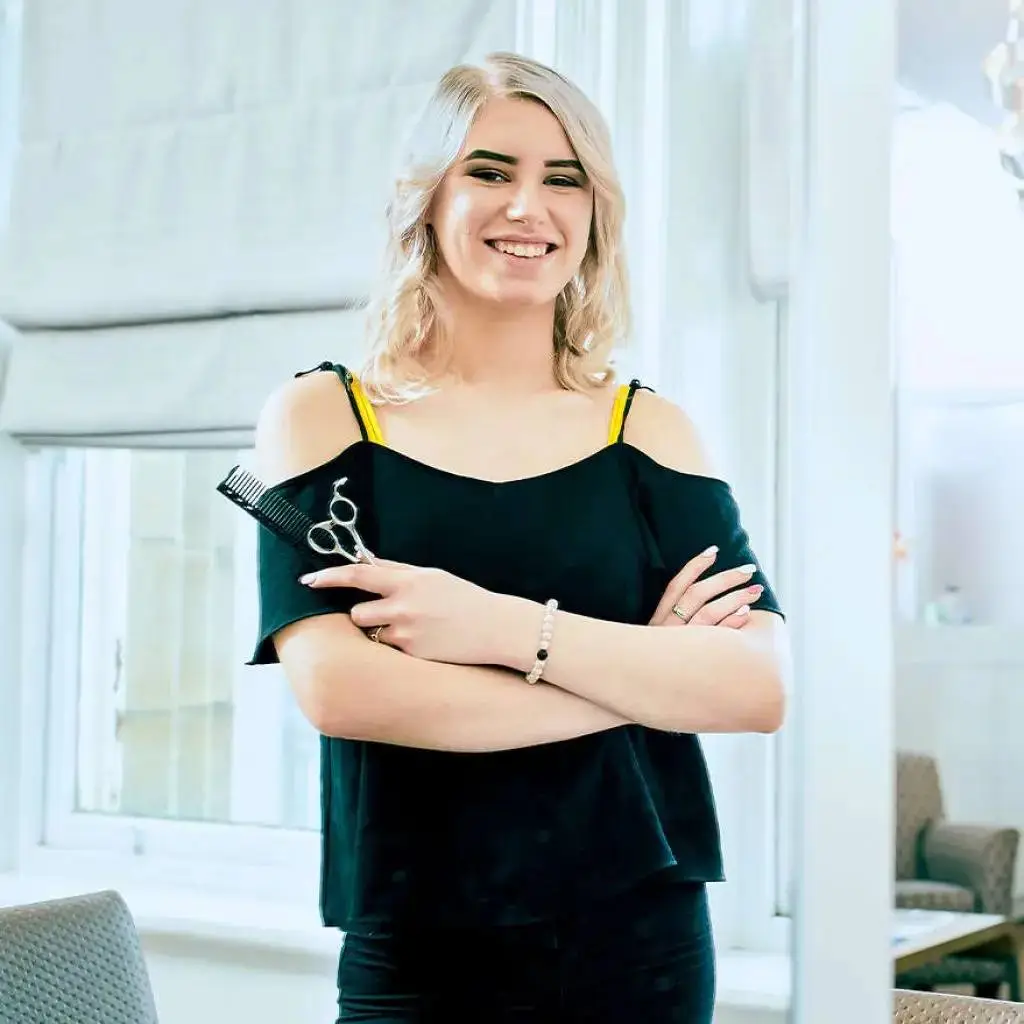 A smiling woman in a black top with scissors and comb, standing confidently in a bright, modern salon setting.