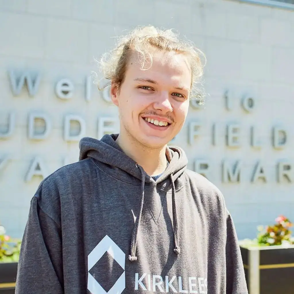 A young man in a grey hoodie smiling in front of a stone wall with partially visible text.