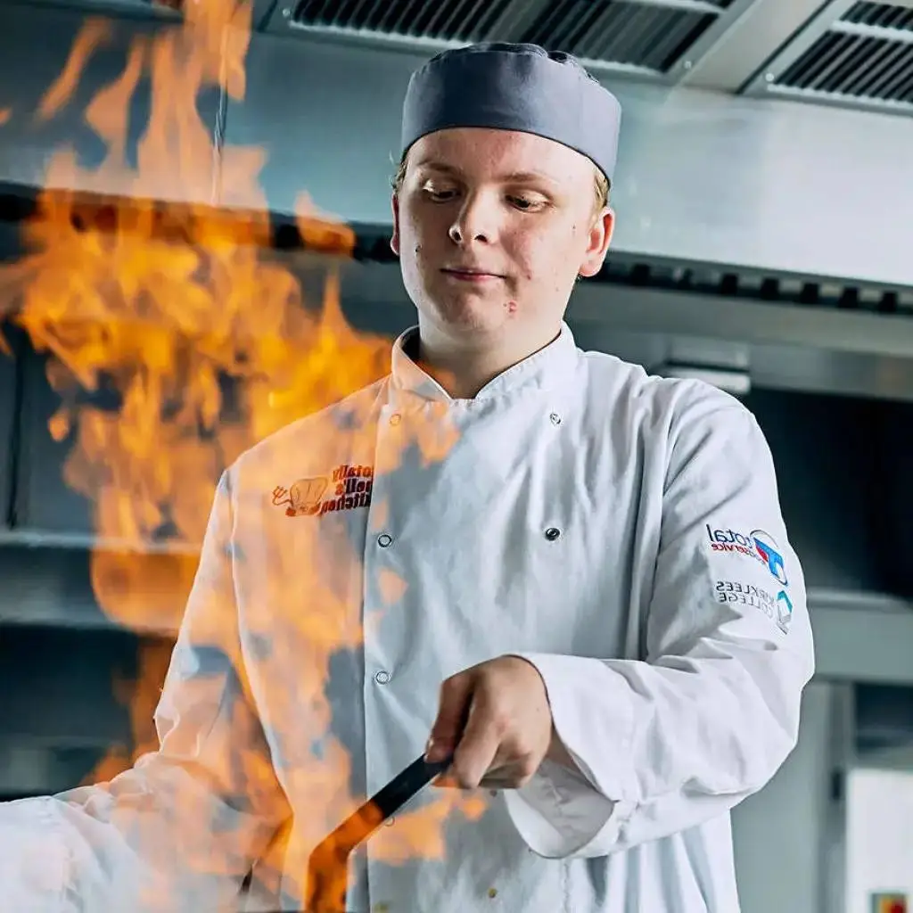 A chef in a white uniform flambéing a dish in a professional kitchen, showcasing culinary skills and technique.