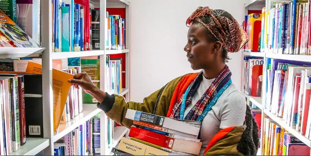 A person in a library, holding books and reaching for a folder on a shelf filled with various books.