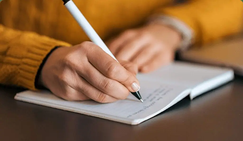 A person in a yellow jumper writing in a notebook with a white pen on a dark table.