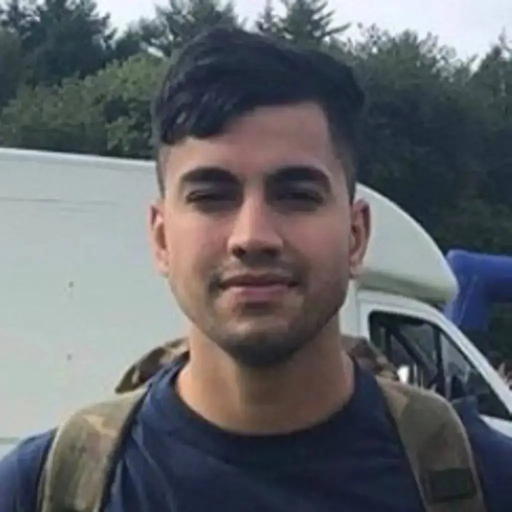 A man with short dark hair and a stubble, wearing a blue t-shirt and a camouflage backpack, stands in front of a white van and trees.