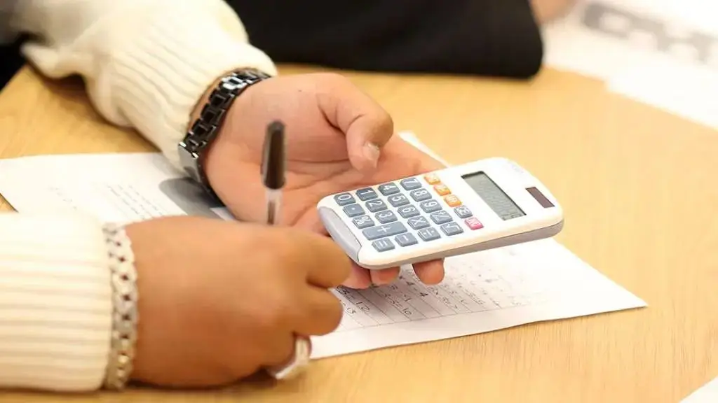 person using a calculator and pen on a worksheet at a wooden desk.