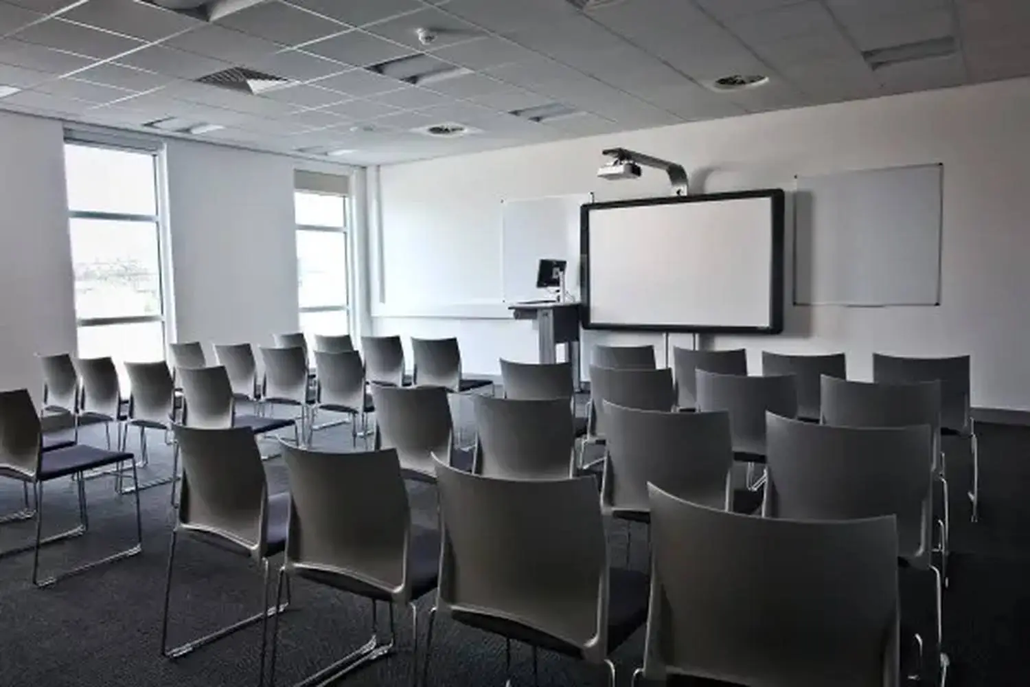A university lecture room with rows of grey chairs facing a projector screen and whiteboard, illuminated by natural light from large windows.