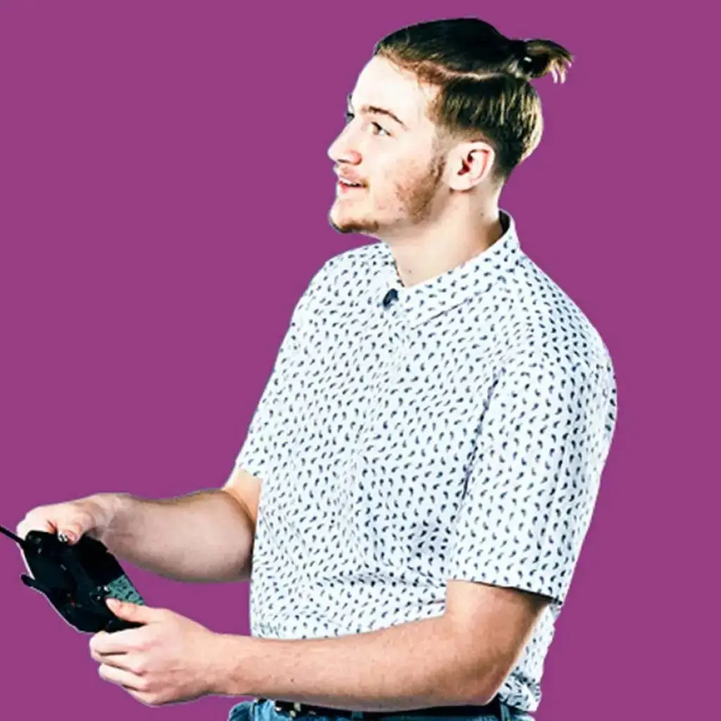 A man with a man bun holding a game controller, smiling against a purple background. Suitable for a gaming or technology education context.