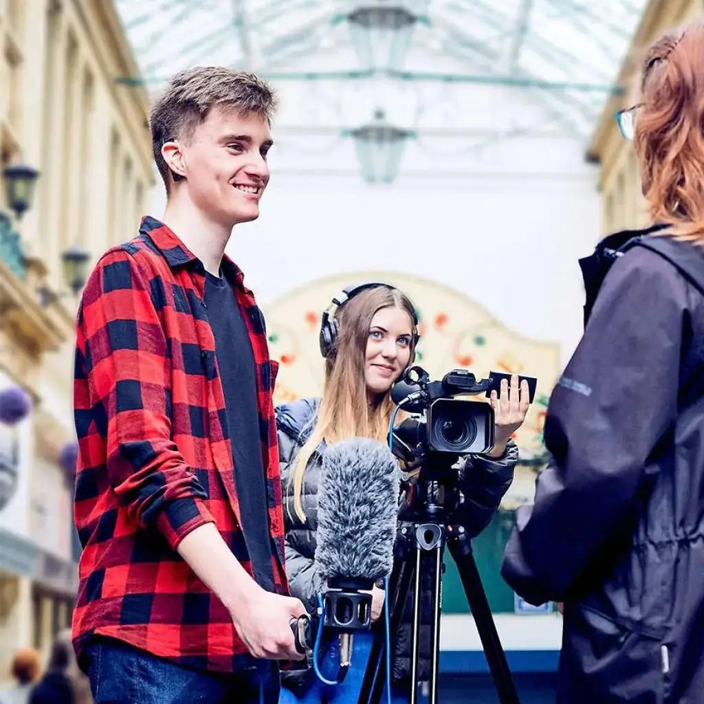 A student in a red checkered shirt being interviewed by a camerawoman in a covered arcade.