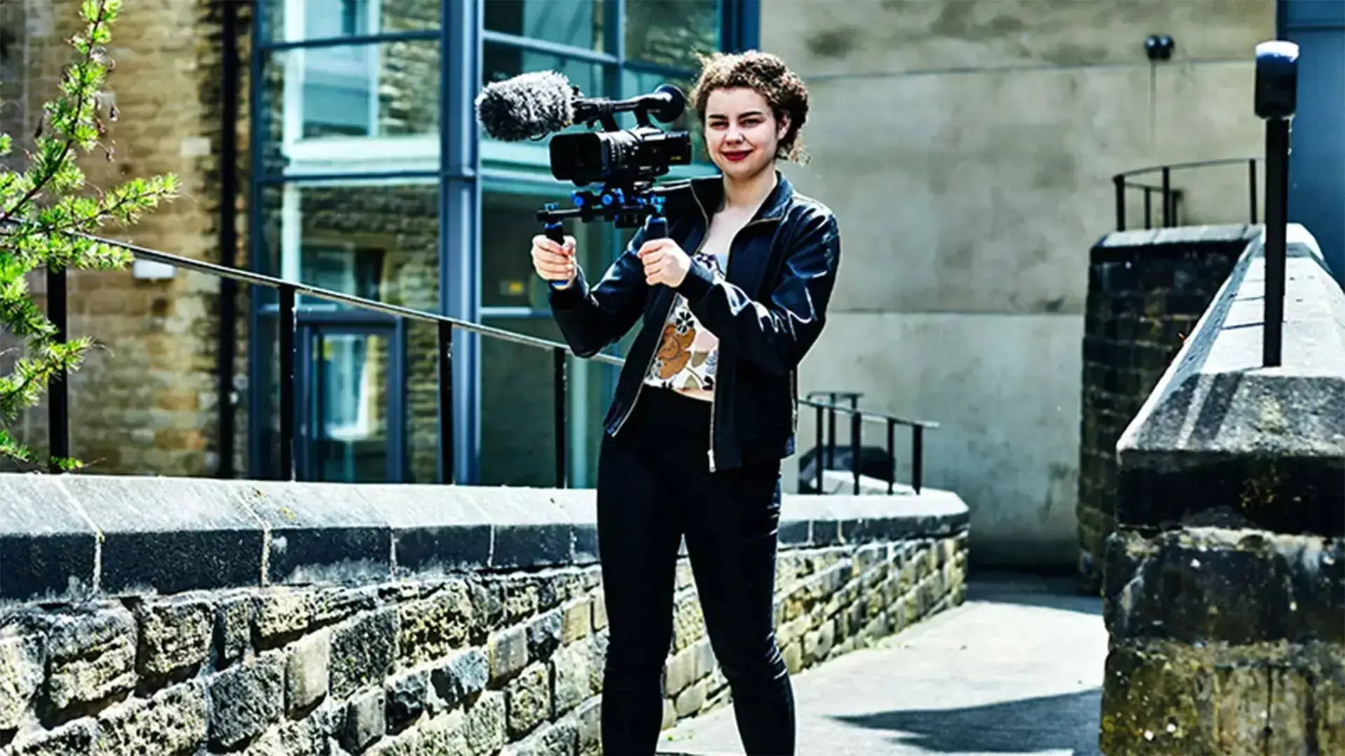 A student in a black tracksuit holding a professional video camera outdoors, smiling at the camera. Modern building in the background.